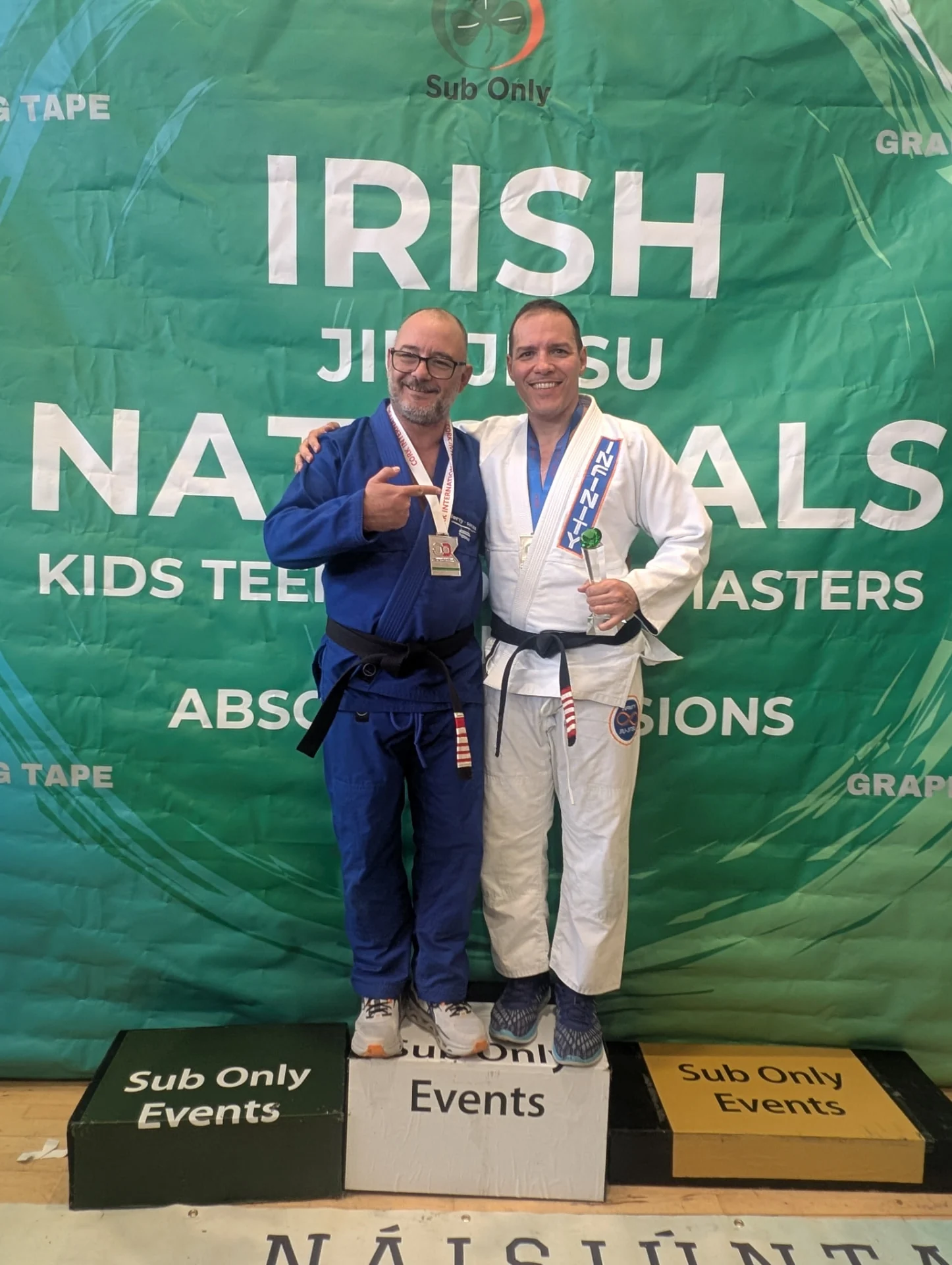 Fabiano on the podium at the Irish Jiu-Jitsu Nationals in his Infinity gi, holding a trophy alongside another black belt