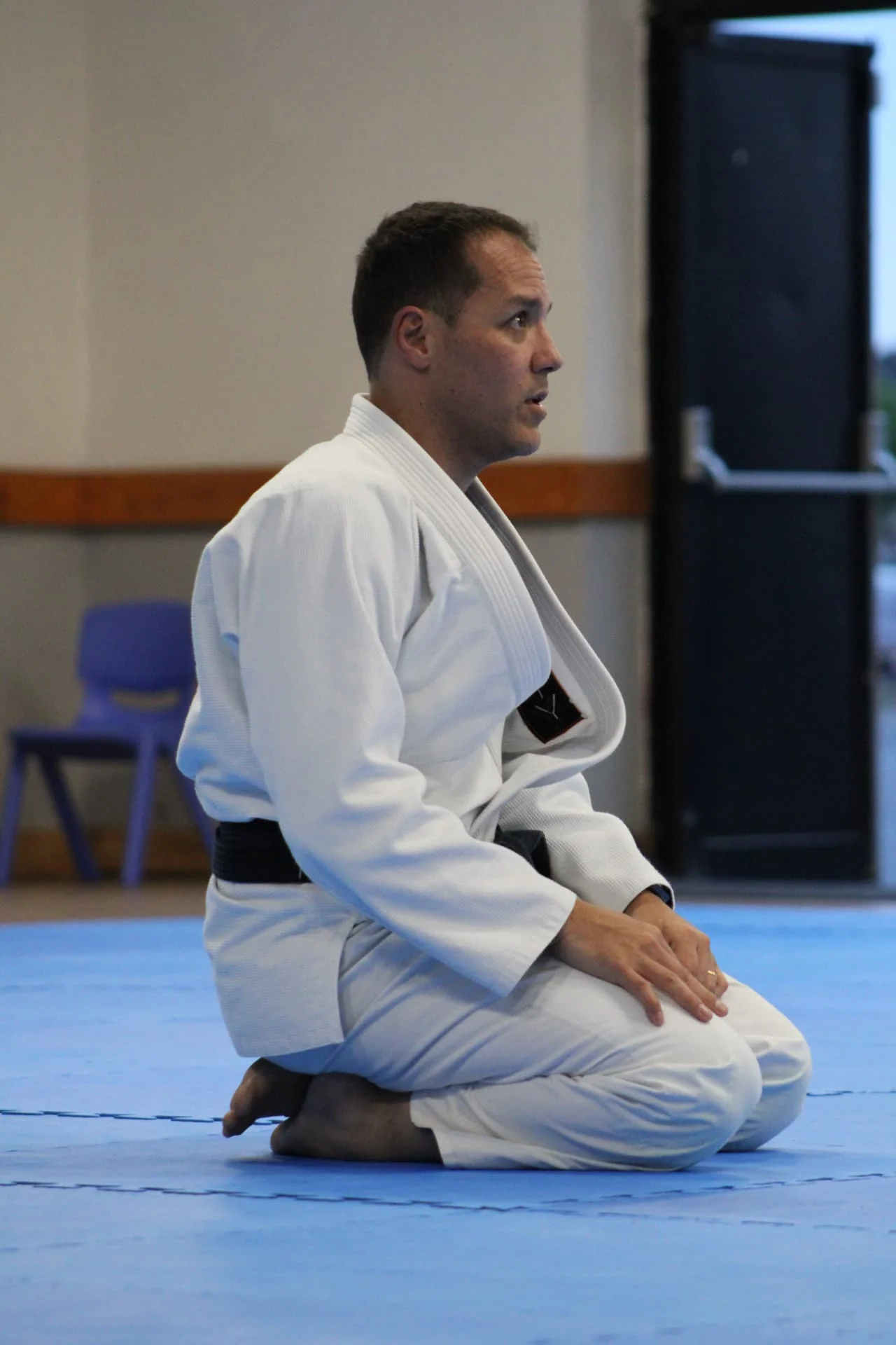 Professor Fabiano kneeling on the mats in a white gi, calm and attentive in a teaching moment at Infinity Jiu-Jitsu