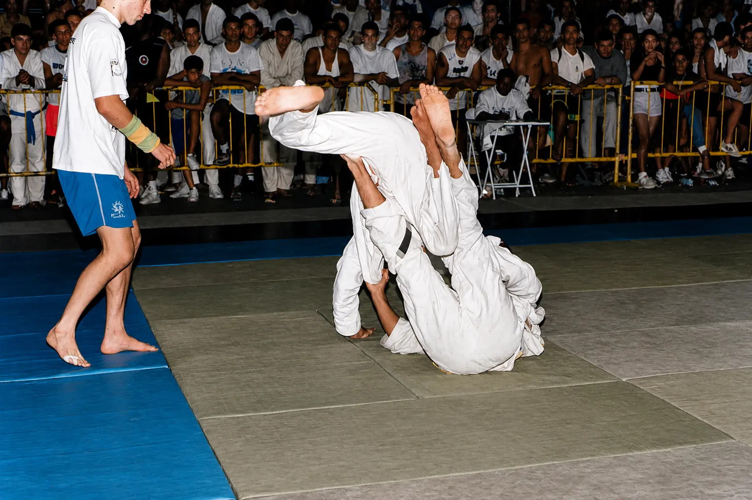 Colour photograph of a dynamic grappling exchange at a Brazilian tournament in front of a large crowd