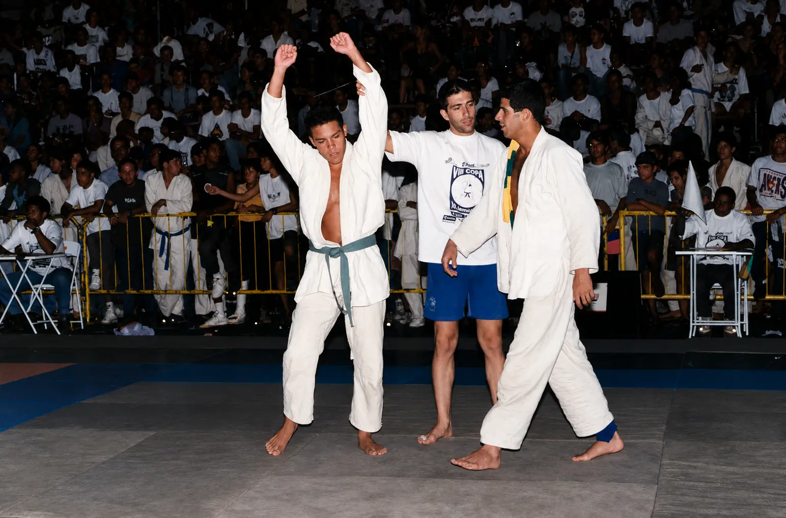 A teenage Fabiano raises both arms in victory on the mat in Brazil, the referee standing between the two competitors in front of a packed crowd
