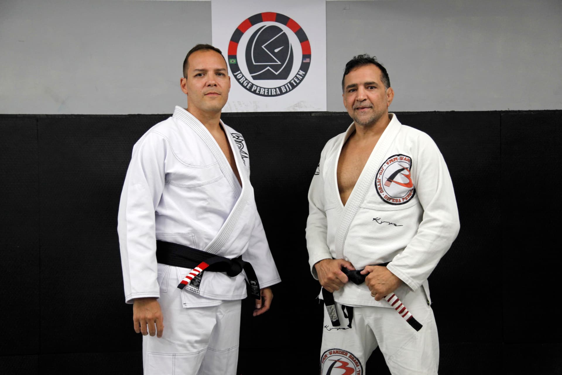 Fabiano standing beside Wander Braga, both in white gis, in front of the academy wall