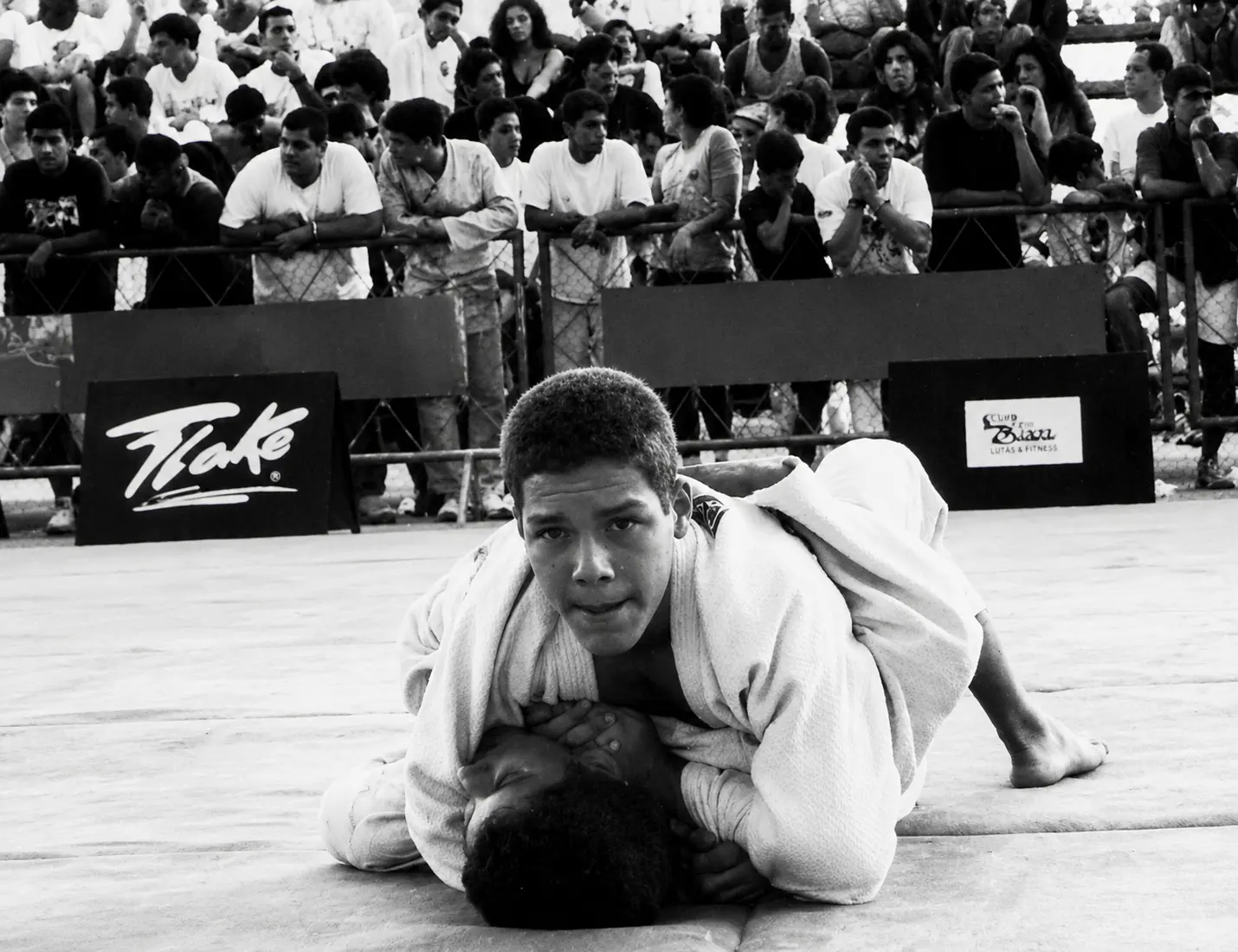 Black and white photograph of a young Fabiano controlling his opponent on the ground during a Brazilian tournament, spectators watching behind the barrier
