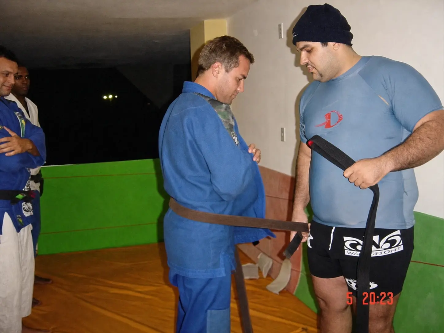 Fabiano standing on the mat as his Brazilian Jiu-Jitsu black belt is presented to him, with teammates watching