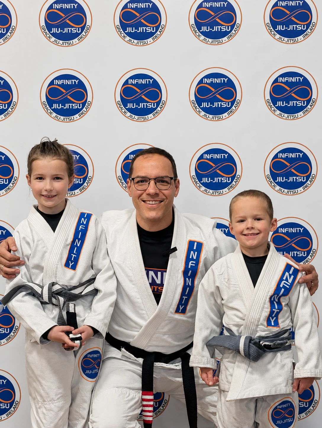 Professor Fabiano with two young students holding their new belts in front of the Infinity Jiu-Jitsu backdrop