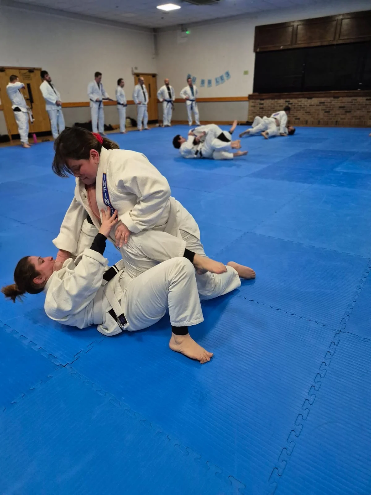Two adult students practising a controlled technique on the mats while the rest of the class trains in the background